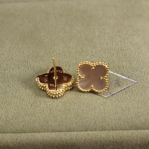 K&ouml;nnte beinhalten: Ein Paar goldfarbene Ohrstecker in Kleeblattform. Jeder Ohrring hat ein vierbl&auml;ttriges Kleeblatt-Design mit einem Perlmutt-Mittelteil und einem Perlenrand. Die Ohrringe haben einen Stecker- und Verschluss.