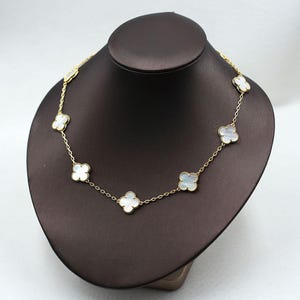 May include: A gold-colored necklace featuring a series of white clover-shaped pendants. The necklace is displayed on a brown jewelry bust, showcasing its delicate design and intricate details. The clover pendants are evenly spaced along the chain.