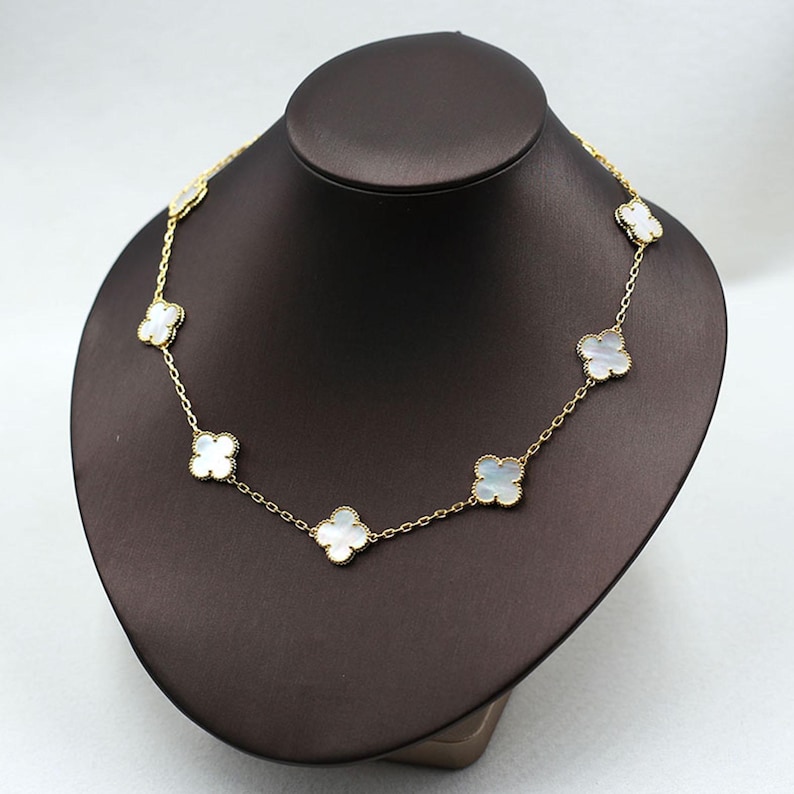 May include: A gold-colored necklace featuring a series of white clover-shaped pendants. The necklace is displayed on a brown jewelry bust, showcasing its delicate design and intricate details. The clover pendants are evenly spaced along the chain.