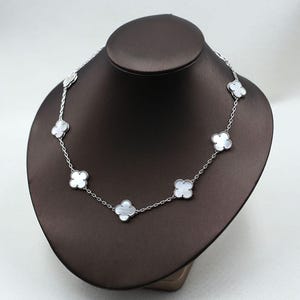 May include: A silver necklace with a delicate chain and clover-shaped pendants. Each pendant features a white, mother-of-pearl-like center. The necklace is displayed on a brown jewelry bust.