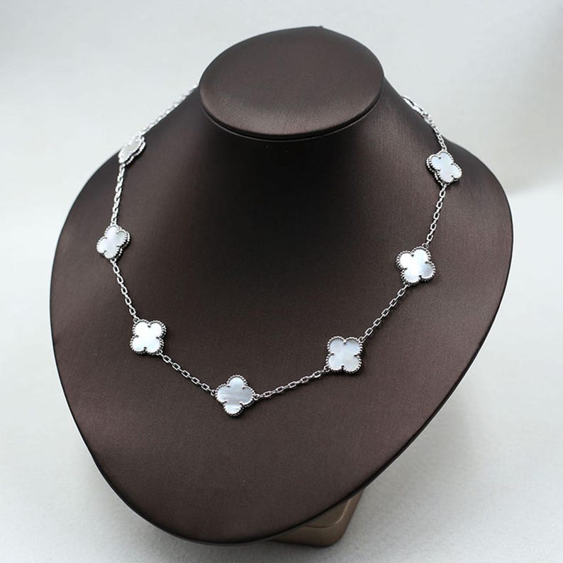 May include: A silver necklace with a delicate chain and clover-shaped pendants. Each pendant features a white, mother-of-pearl-like center. The necklace is displayed on a brown jewelry bust.