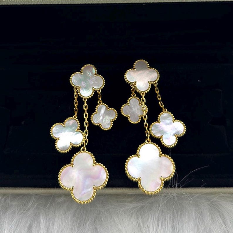 May include: Dangling earrings featuring four-leaf clover designs. Each earring has multiple clover-shaped pendants in white mother-of-pearl, outlined in gold. The pendants are connected by delicate gold chains, creating a layered effect.
