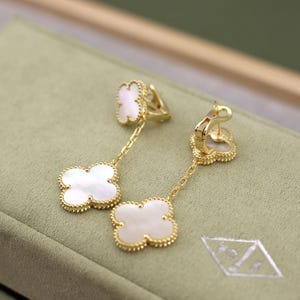 May include: A pair of gold-colored drop earrings featuring a four-leaf clover design. Each clover is white with a gold beaded border. The earrings hang from a gold chain and have a clasp closure.