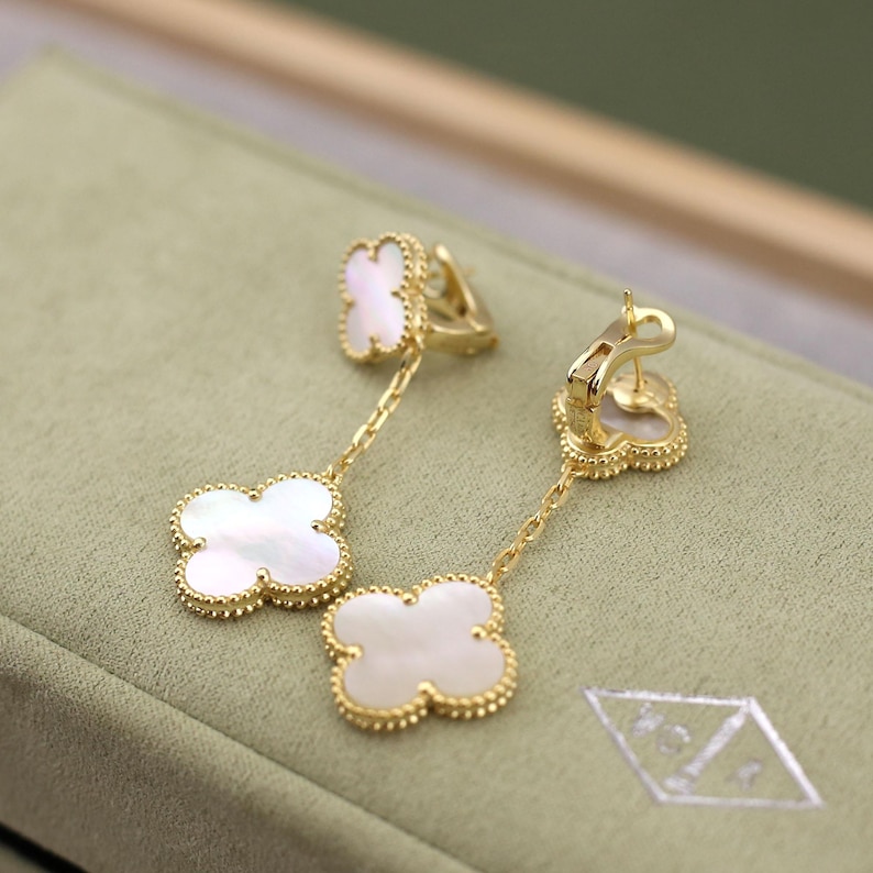 May include: A pair of gold-colored drop earrings featuring a four-leaf clover design. Each clover is white with a gold beaded border. The earrings hang from a gold chain and have a clasp closure.