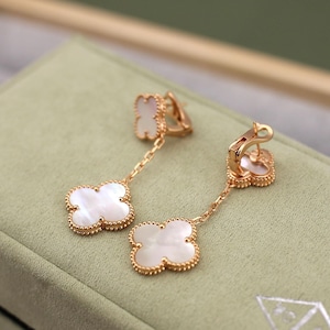 May include: Gold-tone clover-shaped earrings with white mother-of-pearl inserts. The earrings feature a chain and a beaded border around the clover design. The earrings are displayed on a green surface.
