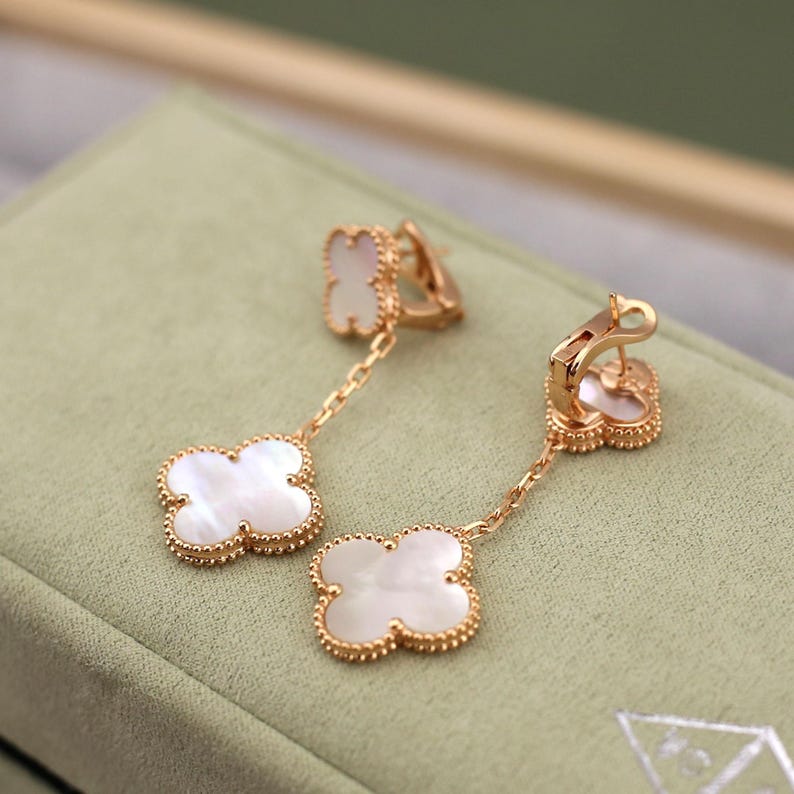 May include: Gold-tone clover-shaped earrings with white mother-of-pearl inserts. The earrings feature a chain and a beaded border around the clover design. The earrings are displayed on a green surface.