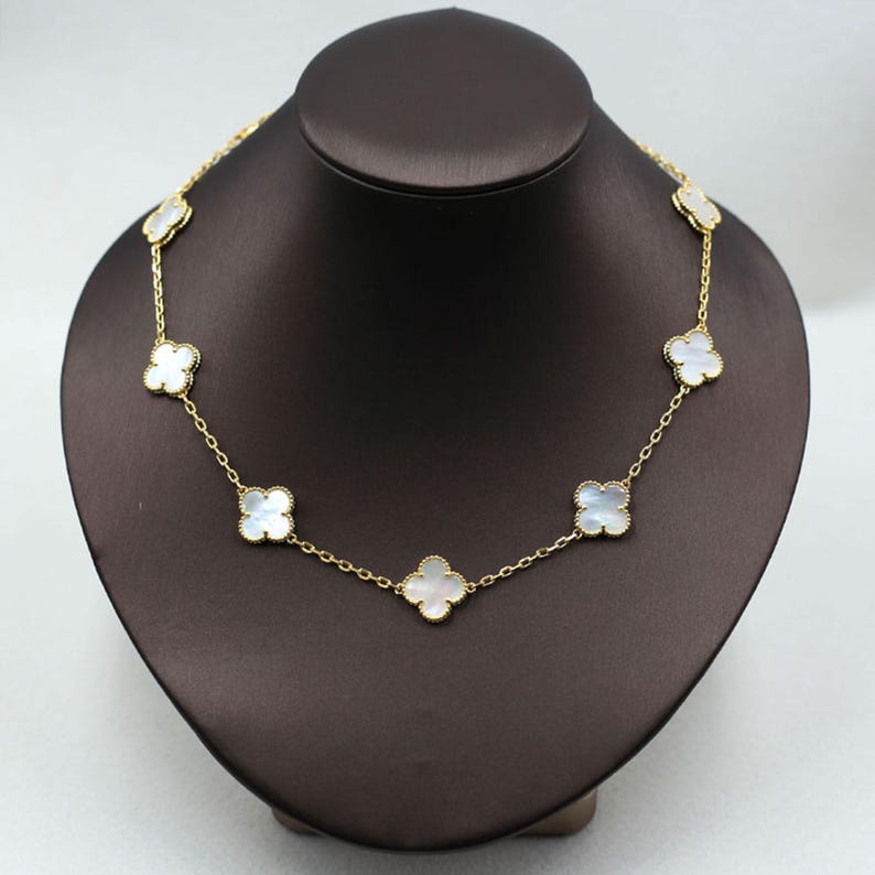 May include: A gold-colored necklace featuring a series of white clover-shaped pendants. The necklace is displayed on a brown jewelry bust. The chain is delicate, and the pendants have a pearlescent sheen.