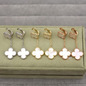 May include: Four pairs of clover-shaped drop earrings. The earrings are in silver, gold, and rose gold tones, each with a white mother-of-pearl clover pendant. The earrings are displayed on a green velvet surface.