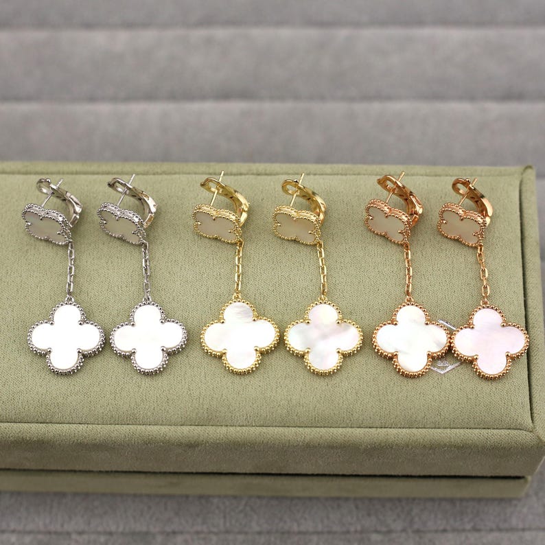 May include: Four pairs of clover-shaped drop earrings. The earrings are in silver, gold, and rose gold tones, each with a white mother-of-pearl clover pendant. The earrings are displayed on a green velvet surface.