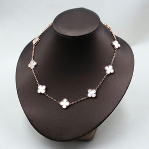 May include: A rose gold necklace featuring a series of white clover-shaped pendants. The necklace is displayed on a black mannequin, showcasing the delicate chain and the lustrous appearance of the clover charms. The background is a neutral gray.