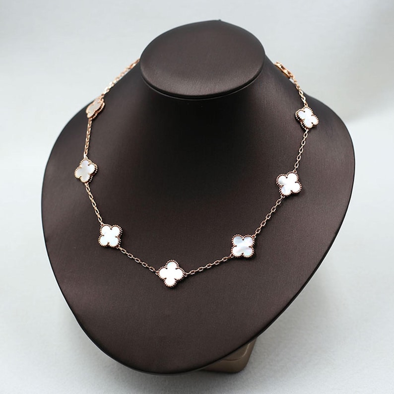 May include: A rose gold necklace featuring a series of white clover-shaped pendants. The necklace is displayed on a black mannequin, showcasing the delicate chain and the lustrous appearance of the clover charms. The background is a neutral gray.