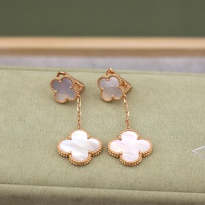 May include: A pair of gold-colored clover-shaped earrings with white mother-of-pearl inlays. The earrings feature a small clover at the top and a larger clover pendant, connected by a delicate chain. The edges are adorned with small gold beads.