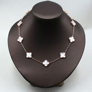 May include: A rose gold necklace featuring seven white clover-shaped pendants. The necklace is displayed on a black jewelry bust. The clover pendants are evenly spaced along the delicate chain, creating a symmetrical design.