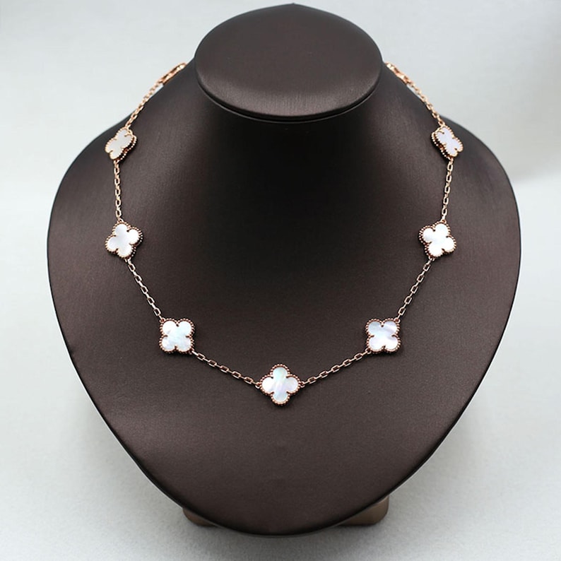 May include: A rose gold necklace featuring seven white clover-shaped pendants. The necklace is displayed on a black jewelry bust. The clover pendants are evenly spaced along the delicate chain, creating a symmetrical design.