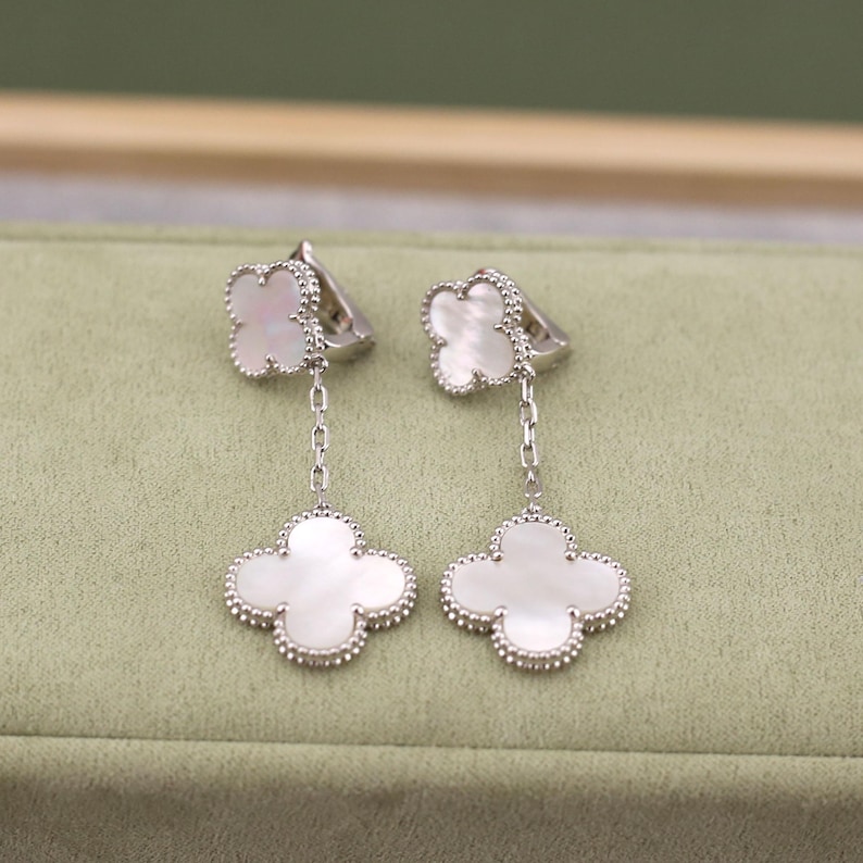 May include: A pair of silver-toned dangle earrings. Each earring features a four-leaf clover design with a white, pearlescent center and a beaded border. The earrings hang from a small, matching clover-shaped stud.