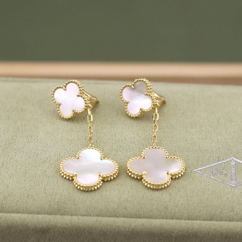 May include: Gold-tone clover-shaped earrings with white mother-of-pearl inlays. The earrings feature a delicate chain connecting the clover to the ear post. The clover design is outlined with a beaded border, adding a touch of elegance.