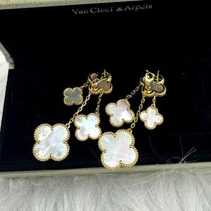 May include: Gold-toned clover-shaped earrings with mother-of-pearl inlays. The earrings feature multiple clover charms of varying sizes, connected by delicate chains. The earrings are displayed in a black jewelry box with the brand name "Van Cleef & Arpels".