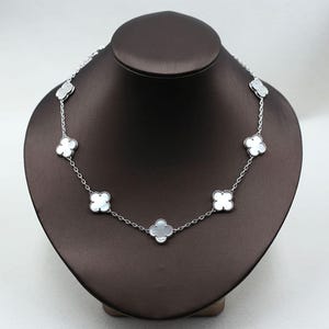 May include: A silver necklace with white clover-shaped pendants displayed on a brown jewelry bust. The necklace has a delicate chain and the clover pendants are evenly spaced along the chain.