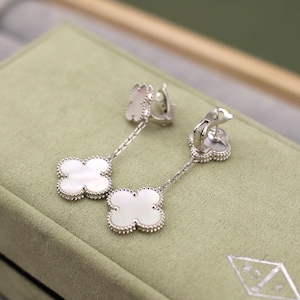 May include: A pair of silver-toned drop earrings featuring a four-leaf clover design with a white mother-of-pearl inlay. The earrings have a beaded border and are displayed on a light green surface.