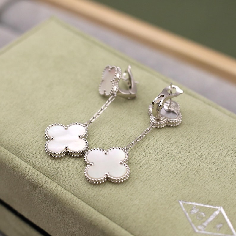 May include: A pair of silver-toned drop earrings featuring a four-leaf clover design with a white mother-of-pearl inlay. The earrings have a beaded border and are displayed on a light green surface.
