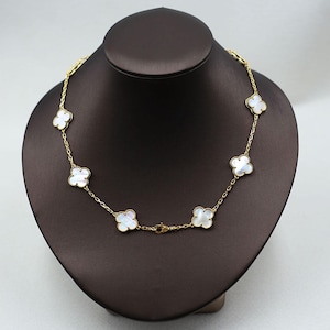May include: A gold-colored necklace with seven white clover-shaped pendants. The necklace is displayed on a brown jewelry bust. The chain is delicate, and the clasps are visible.
