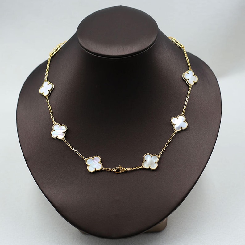 May include: A gold-colored necklace with seven white clover-shaped pendants. The necklace is displayed on a brown jewelry bust. The chain is delicate, and the clasps are visible.