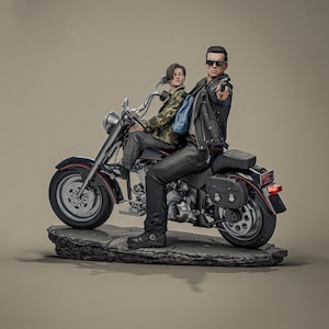 May include: A detailed figurine of two figures on a black motorcycle. One figure, wearing sunglasses and a black leather jacket, points a gun. The other figure wears a camouflage jacket and sits behind the first. The motorcycle is on a stone base.