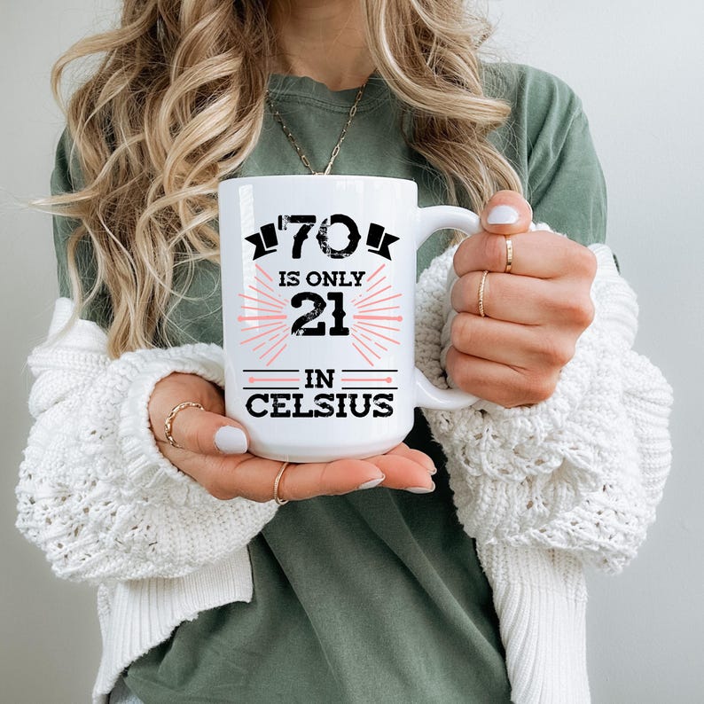 以下が含まれることがあります： 黒とピンクで「70 is only 21 in Celsius」と書かれた白いセラミックマグ。マグには大きなハンドルがあり、白いセーターを着た人が持っています。