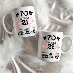 以下が含まれることがあります： 「70 is only 21 in Celsius」の文字が黒とピンクでプリントされた、白いセラミックマグカップが2つ。マグカップには取っ手があり、白い羽を背景に配置されています。