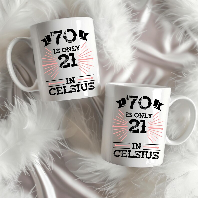 以下が含まれることがあります： 「70 is only 21 in Celsius」の文字が黒とピンクでプリントされた、白いセラミックマグカップが2つ。マグカップには取っ手があり、白い羽を背景に配置されています。