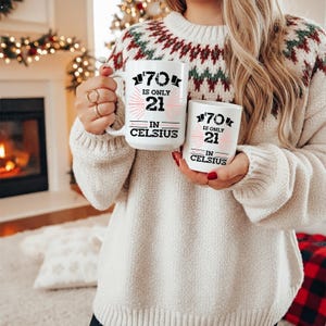 以下が含まれることがあります： 黒とピンクで「70 is only 21 in Celsius」の文字が入った、大きいのと小さいの2つの白いセラミックマグ。クリーム色のセーターを着た人がマグカップを持っています。背景には暖炉とクリスマスの飾りがあります。