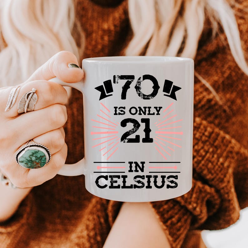 以下が含まれることがあります： 「70 is only 21 in Celsius」の文字が黒とピンクでプリントされた白いセラミックマグカップ。指に指輪をした人がマグカップを持っています。