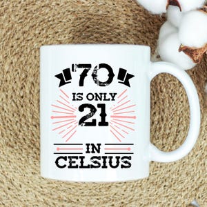 以下が含まれることがあります： 白のセラミックマグカップ。黒とピンクで「70 is only 21 in Celsius」の文字がプリントされています。マグカップにはカーブしたハンドルが付いており、温かい飲み物に使用される可能性があります。デザインはマグカップの中央に配置されています。
