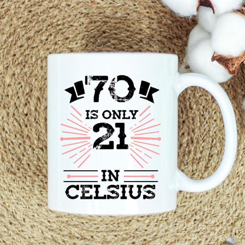 以下が含まれることがあります： 白のセラミックマグカップ。黒とピンクで「70 is only 21 in Celsius」の文字がプリントされています。マグカップにはカーブしたハンドルが付いており、温かい飲み物に使用される可能性があります。デザインはマグカップの中央に配置されています。