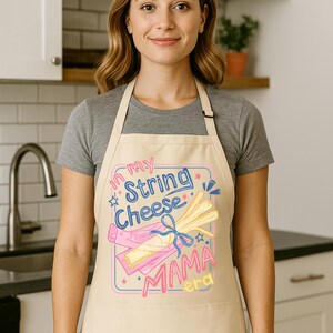 May include: Beige apron with a graphic that reads "In my String Cheese Mama era." The graphic features illustrations of string cheese and a pink and blue design. The apron is worn by a person in a kitchen setting.