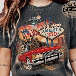 May include: A charcoal grey t-shirt featuring a graphic print of a red convertible car driven by Bigfoot, with an alien and a moth-like creature as passengers. Above the car is a UFO, an American flag, and a sign that reads Welcome America 250.