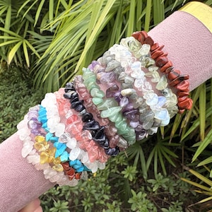 May include: A collection of gemstone chip bracelets in various colors, including blue, purple, green, and red, are displayed on a pink cylindrical stand with a gold rim. The bracelets are made of small, irregular-shaped stones.