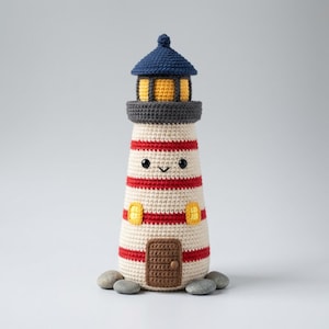 May include: A crocheted lighthouse toy featuring a cream-colored body with horizontal red stripes, a dark blue roof, and a yellow lantern room. The structure includes a brown door, two yellow windows, and a smiling face with black bead eyes.