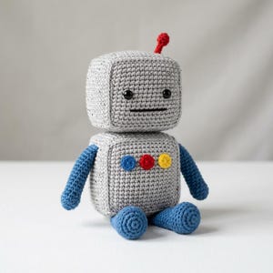 May include: A crocheted robot toy with a square grey head and body, blue arms and legs, and a small red antenna. The face features two black bead eyes and a straight black stitched mouth. Three small circular buttons in blue, red, and yellow are attached to the chest.