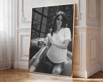May include: A black and white framed wall art print featuring a person with long dark hair wearing a short-sleeved white t-shirt and shorts, standing next to the side of a car. The artwork is displayed in a thin gold frame.