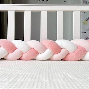 May include: A pink and white braided crib bumper. The bumper is made of soft, plush material and is designed to protect the baby from the crib's hard surfaces. The bumper is placed inside a white cot.