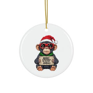 May include: A circular Christmas decoration with a chimpanzee wearing a Santa hat and sunglasses. The chimpanzee holds a sign that reads "MERRY CHRISTMAS YA FILTHY ANIMALS". The ornament has a gold hanging string.