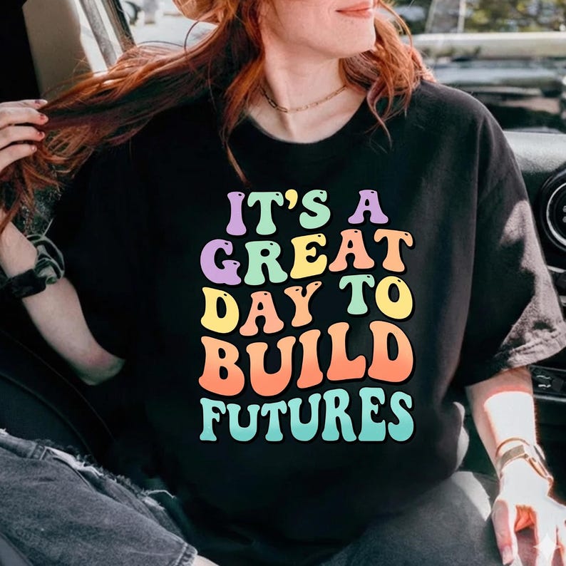 Puede incluir: Camiseta negra con el texto "IT'S A GREAT DAY TO BUILD FUTURES" en letras coloridas estilo burbuja. El texto est&aacute; en tonos morado, verde, amarillo, naranja y azul. Una prenda casual.