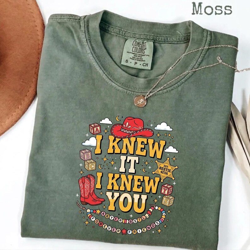 May include: A moss green crew neck t-shirt featuring a graphic print with the text I KNEW IT I KNEW YOU. The design includes a red cowboy hat, red cowboy boots, alphabet blocks, a sheriff star badge, and a beaded necklace with the text BESTIE 4EVA.