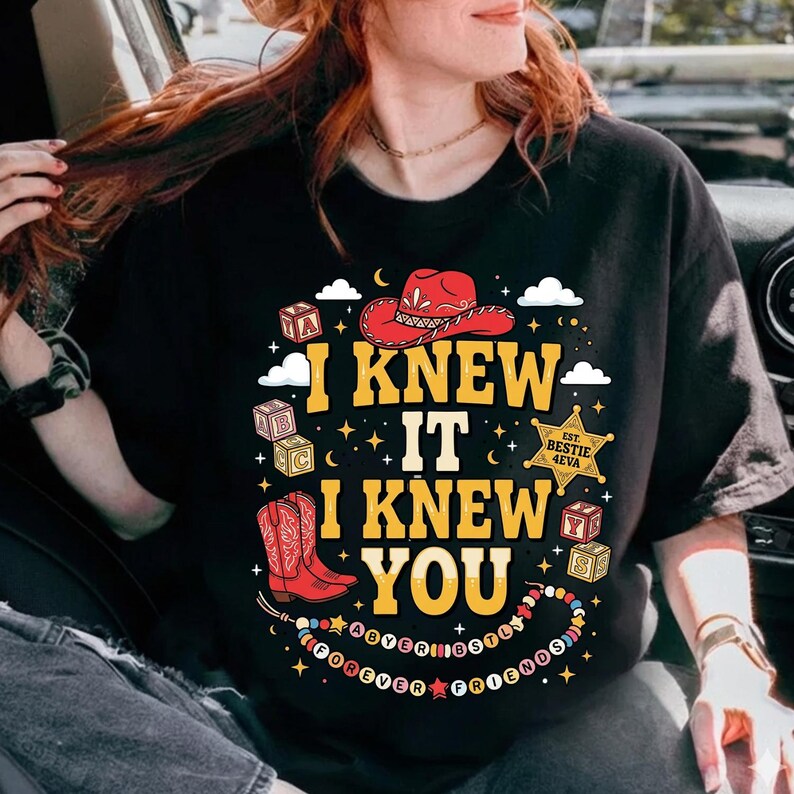 May include: A black t-shirt featuring a graphic print with the text I KNEW IT I KNEW YOU. The design includes a red cowboy hat, red cowboy boots, alphabet blocks, a gold star badge labeled EST. BESTIE 4EVA, and a beaded necklace with the text FOREVER FRIENDS.
