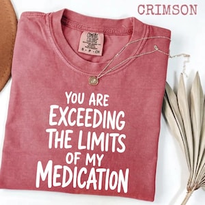 May include: A crimson short-sleeved t-shirt with a crew neckline and a white graphic print that reads, "YOU ARE EXCEEDING THE LIMITS OF MY MEDICATION." The shirt features a Comfort Colors brand label at the inner neck and a visible thin gold chain.