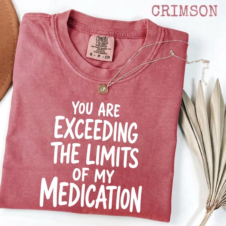 May include: A crimson short-sleeved t-shirt with a crew neckline and a white graphic print that reads, "YOU ARE EXCEEDING THE LIMITS OF MY MEDICATION." The shirt features a Comfort Colors brand label at the inner neck and a visible thin gold chain.