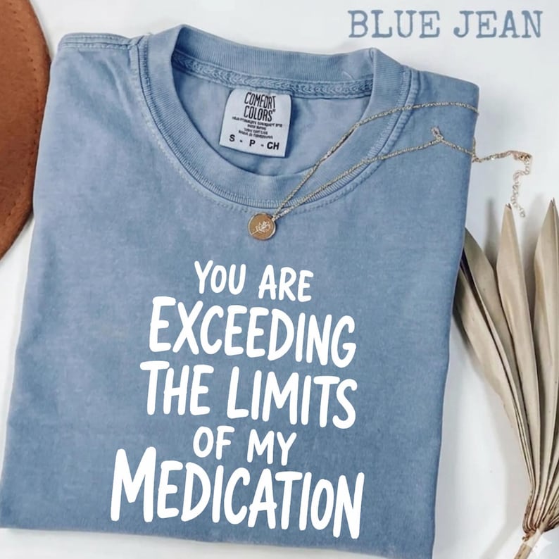 May include: A blue jean colored t-shirt with a crew neckline and a Comfort Colors brand label at the inner collar. The shirt features white text that reads: YOU ARE EXCEEDING THE LIMITS OF MY MEDICATION. A thin gold chain necklace rests on the fabric.