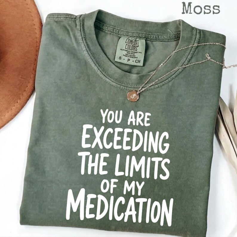 May include: A moss green short-sleeve t-shirt with a crew neckline. The shirt features white printed text that reads, "YOU ARE EXCEEDING THE LIMITS OF MY MEDICATION." A thin gold-toned chain necklace with a small circular pendant rests on the fabric.