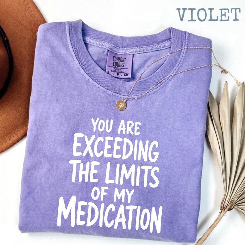 May include: A violet short-sleeved t-shirt with a crew neckline and a Comfort Colors brand label. The shirt features white text printed on the front that reads, "YOU ARE EXCEEDING THE LIMITS OF MY MEDICATION" in a bold, sans-serif font style.